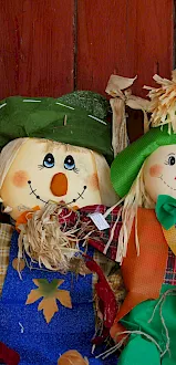 A metal container sits beside two straw scarecrow dolls wearing green hats and plaid clothes, decorated against a red wooden wall.