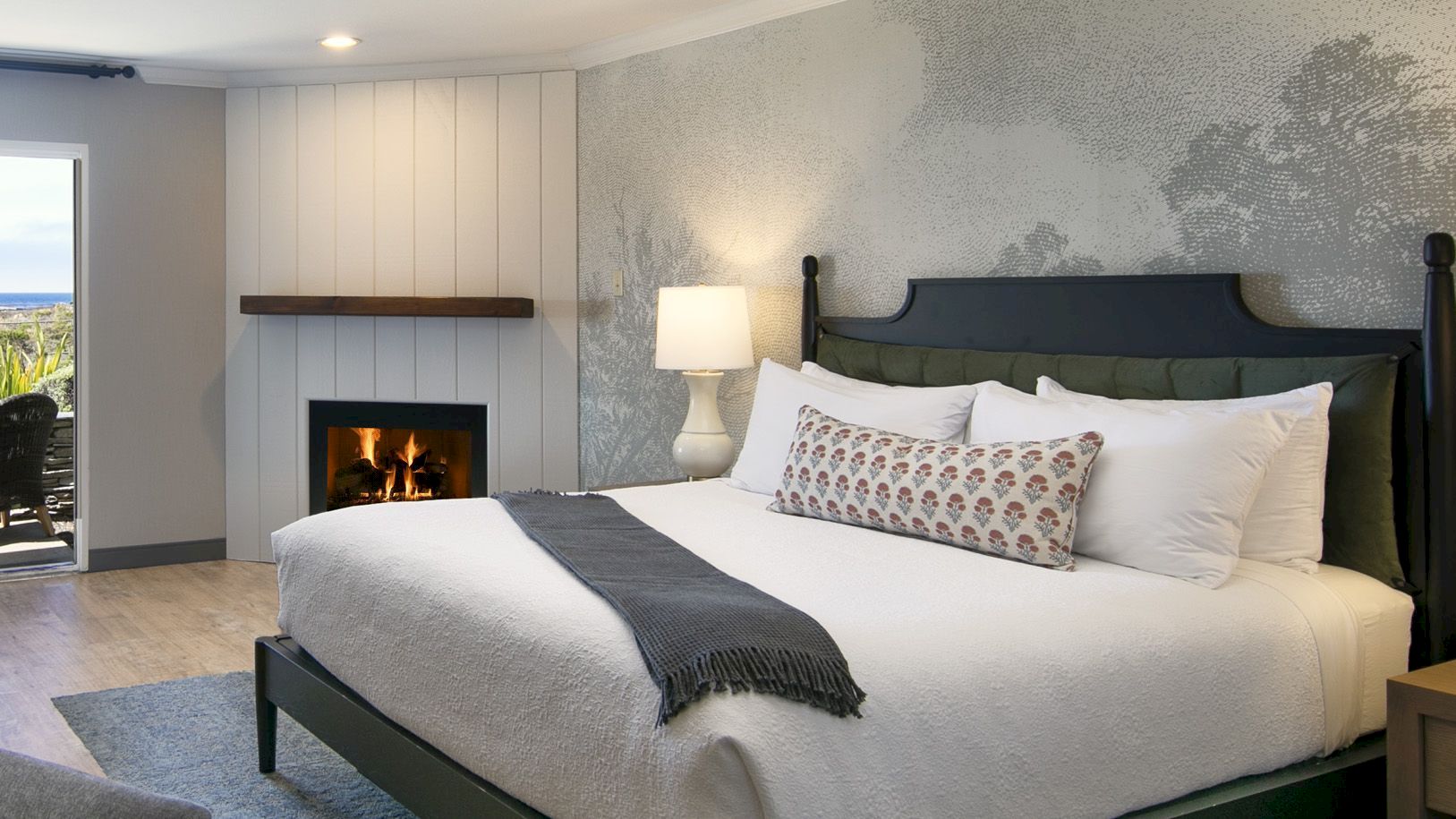 A modern bedroom with a large bed, two bedside lamps, and a wall-mounted fireplace, with curtains and an ocean-view window.