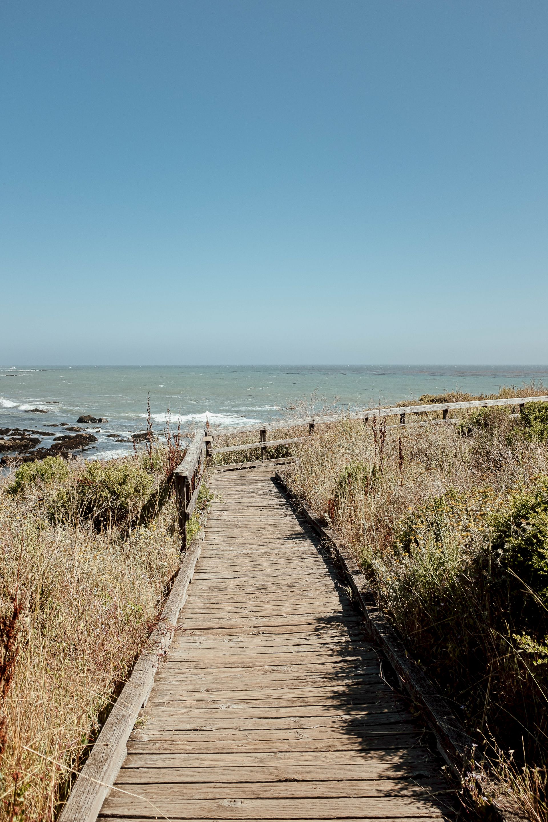 A wooden boardwalk through sandy dunes leads to a choppy ocean under a wide blue sky.