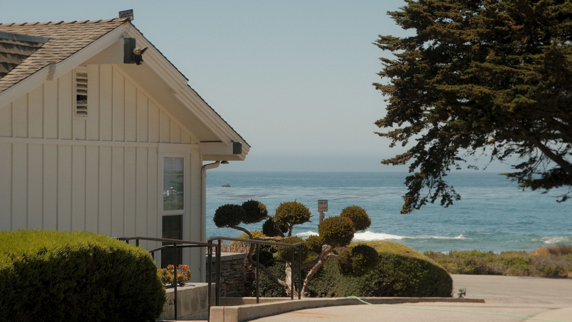 White house beside landscaped bushes overlooks the blue ocean with waves, framed by a large tree at right.