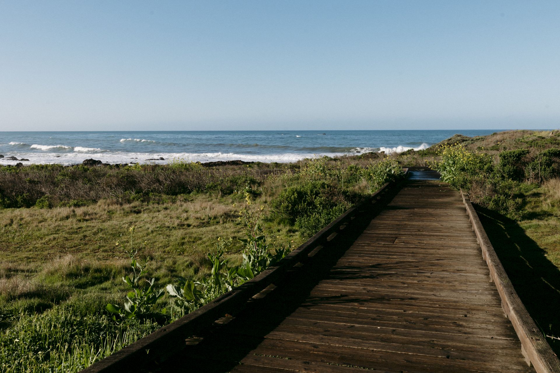 A wooden boardwalk winds through grassy coastal terrain toward the ocean, with waves breaking on the shore under a clear blue sky.