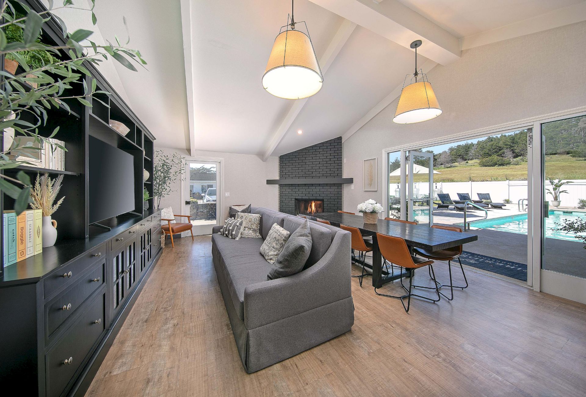 A bright modern living room with a gray sofa, dining table, two pendant lights, and a fireplace opening to a pool terrace.