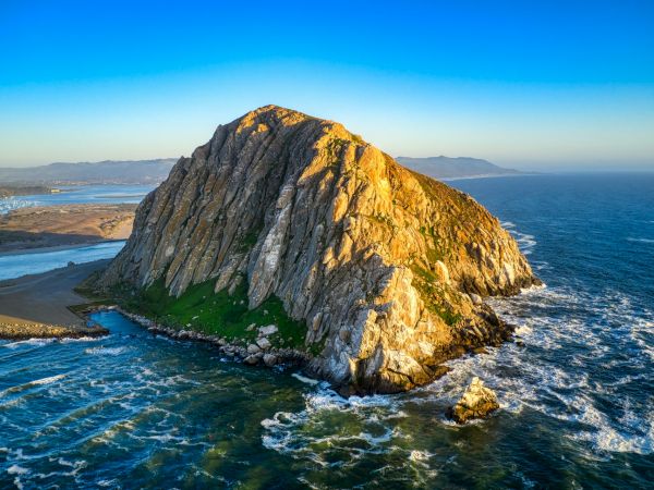 A jagged rocky island rises from blue waves, with sunlight lighting its cliff faces; a sandy shore and distant land stretch into the horizon.