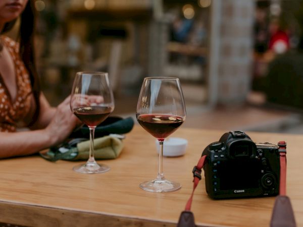 Two wine glasses on a wooden table, a camera nearby, and a blurred person in the background&mdash;romantic outdoor scene.