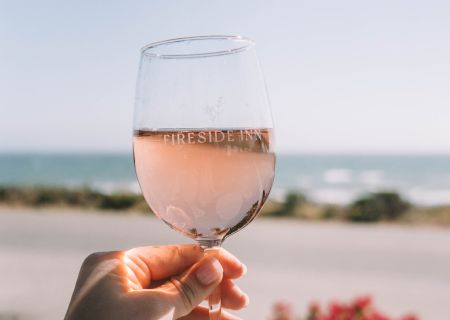 A hand holding a glass of pink wine up against a sunny beach scene with a calm sea in the background, vibes of vacation and sunset.