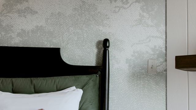 A close-up of a bed with a dark wood frame and green tufted headboard against a white and gray wallpaper.