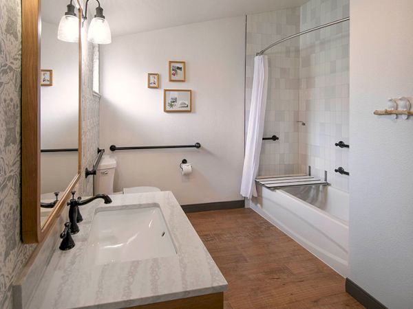A bright bathroom with a double vanity and marble countertop, wall mirror, light fixtures, a bathtub with a glass shower door, towel rails, and wood-look flooring.