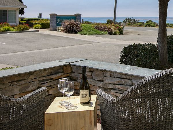 An outdoor patio by the ocean with a stone wall, two wine glasses, and a bottle of Tolosa Chardonnay on a table.
