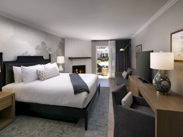 A modern hotel room with a neatly made bed, lamps, a fireplace, seating, and large curtains by the window.