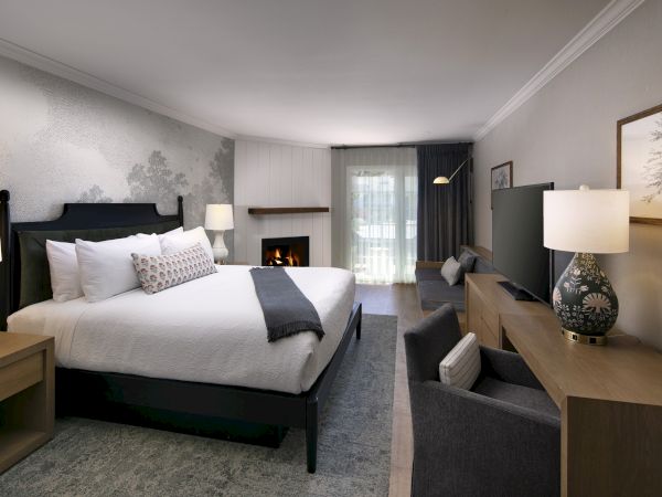 A modern bedroom with a made bed, lamps, a lit fireplace, armchairs, and framed art on textured walls.