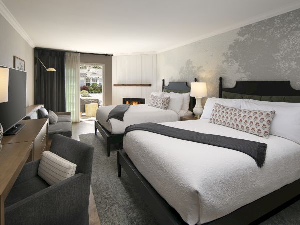 A modern hotel bedroom with two beds, patterned pillows, bedside lamps, a small wall fireplace, and dark curtains by a window.