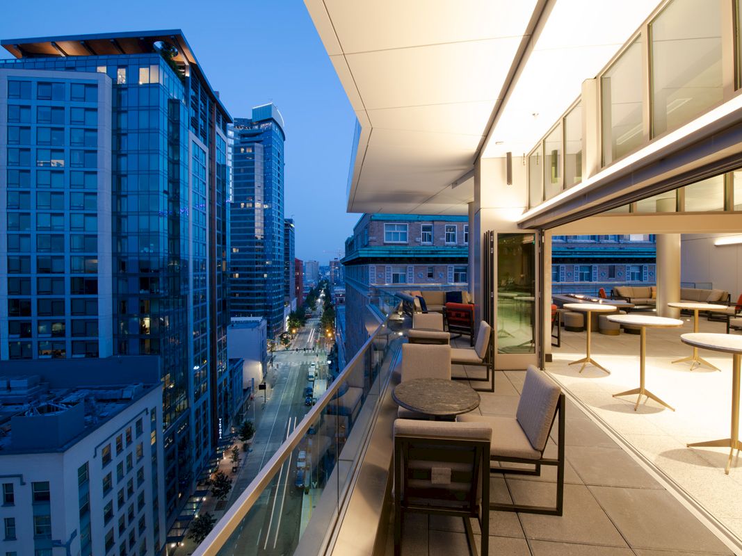 A modern high-rise rooftop terrace with lounge seating overlooks a downtown city street lined with tall buildings.